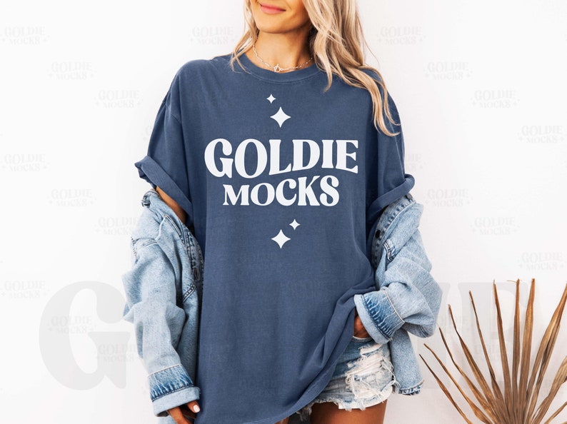 May include: A navy blue t-shirt with the text "GOLDIE MOCKS" in white, along with two white starburst graphics. The shirt is worn with a light blue denim jacket and denim shorts.