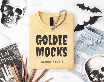 May include: A yellow t-shirt with the text "GOLDIE MOCKS" in black, and a silver chain necklace. The image includes a skull, a palm reading book, and black bat cutouts, along with a pair of denim shorts.