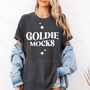 May include: A dark gray t-shirt with the text "GOLDIE MOCKS" in white, along with two white starburst graphics. The shirt is worn with a light blue denim jacket. The model is wearing a silver necklace.