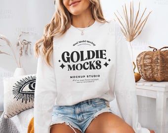May include: White sweatshirt with a black graphic design that says "GOLDIE MOCKS MOCKUP STUDIO" with a star design on either side of the text. The model is wearing denim shorts.