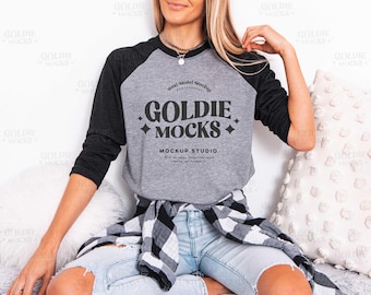 May include: A gray raglan-style t-shirt with black sleeves. The shirt features the text "GOLDIE MOCKS" in a bold, stylized font. The shirt is paired with light blue jeans and a black and white plaid shirt tied around the waist.