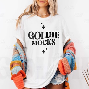 May include: White t-shirt with black text that reads "Goldie Mocks" and small black stars. The shirt is being worn by a person with long blonde hair and a colorful cardigan.