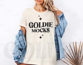 Op de afbeelding: Een wit T-shirt met een zwarte afbeelding waarop "Goldie Mocks" staat met sterren rond de tekst.