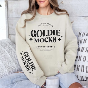 Puede incluir: Una sudadera beige con el texto "Real POD Mockup Photography" y "GOLDIE MOCKS" en negro. El texto "MOCKUP STUDIO" también está en negro y el texto "All of our images feature real people wearing real products" está en negro más pequeño. La sudadera tiene un gráfico en blanco y negro "GOLDIE MOCKS" en la manga izquierda.