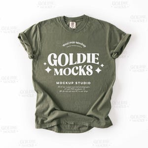 以下が含まれることがあります： オリーブグリーンのTシャツ、袖はロールアップされています。「GOLDIE MOCKS」の文字が白のスタイリッシュなフォントで表示され、「MOCKUP STUDIO」などの文字も表示されています。Tシャツはモックアップです。