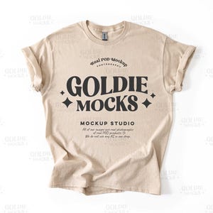 Puede incluir: Una camiseta beige con un gráfico negro que dice "Goldie Mocks" y "Real POD Mockup Photography". El texto también dice "Mockup Studio" y "Todas nuestras imágenes son fotografías reales de productos POD reales. No usamos ninguna IA en nuestra tienda."