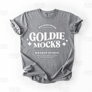Op de afbeelding: Een grijs T-shirt met een wit grafisch ontwerp met de tekst "GOLDIE MOCKS" en "MOCKUP STUDIO".