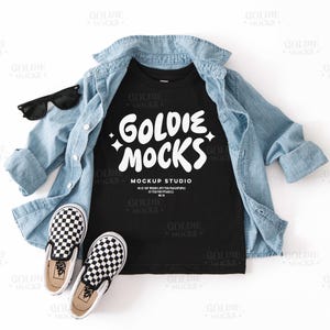 Puede incluir: Camiseta negra con texto blanco que dice "Goldie Mocks Mockup Studio". La camiseta está sobre una superficie blanca con una camisa vaquera azul y zapatos negros y blancos de tela.