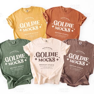 May include: Five different colored t-shirts with the text "Goldie Mocks" and "Mockup Studio" printed on them. The shirts are laid out on a white surface.