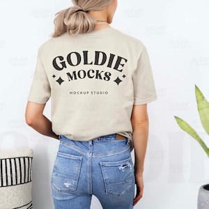 May include: A beige t-shirt with the text "Goldie Mocks" and "Mockup Studio" printed on the back. The shirt is being worn by a person with long blonde hair and blue jeans.
