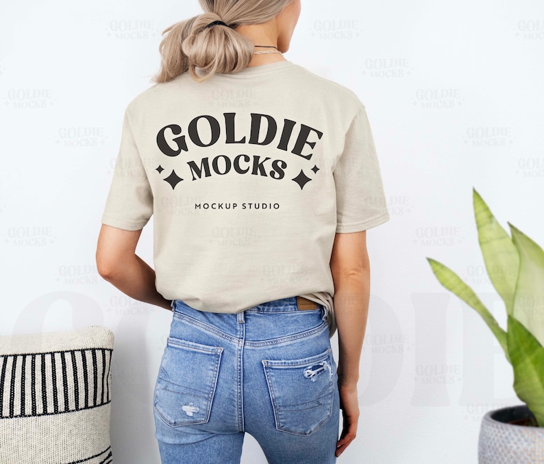 May include: A beige t-shirt with the text "Goldie Mocks" and "Mockup Studio" printed on the back. The shirt is being worn by a person with long blonde hair and blue jeans.