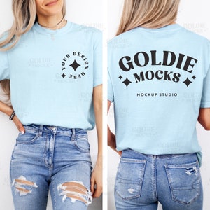 以下が含まれることがあります： 短い袖の薄い青色のTシャツで、前面には「Your Design Here」という文字と星が描かれた丸いデザインがあります。Tシャツの背面には、「Goldie Mocks」という文字と両側に星が2つ、その下に「Mockup Studio」という文字があります。Tシャツは、青いジーンズを履いた人が着用しています。