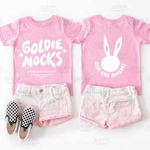 以下が含まれることがあります： ピンクの幼児用Tシャツのモックアップ。背面には白いウサギのグラフィック、前面には「Goldie Mocks Mockup Studio」のテキストがプリントされています。Tシャツはピンクのデニムショーツと白黒のチェック柄のスリッポンシューズと組み合わせています。