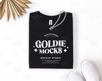 Könnte beinhalten: Ein schwarzes T-Shirt mit dem weißen Text "GOLDIE MOCKS" und einer silbernen Kette. Das Shirt liegt flach auf einer weißen Oberfläche, mit einem getrockneten Palmblatt und weißen Turnschuhen im Hintergrund.
