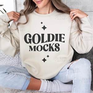 Puede incluir: Una mujer lleva una sudadera de color crema con el texto "GOLDIE MOCKS" en letras negras. La sudadera tiene un corte holgado y se combina con unos vaqueros rotos.