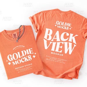 Pode incluir: Duas camisetas laranja com texto branco. Uma camisa exibe "GOLDIE MOCKS MOCKUP STUDIO" e a outra diz "BACK VIEW MOCKUP". Um colar de corrente prateada repousa sobre a camisa dobrada. As camisas estão em um fundo branco.