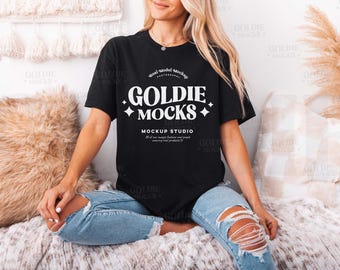 May include: A woman wearing a black t-shirt with the text "Goldie Mocks Mockup Studio" in white. The shirt is a relaxed fit and has a crew neck. She is sitting on a white and brown fuzzy surface.