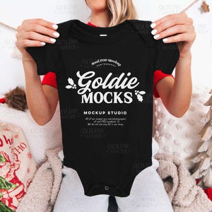 May include: Black baby bodysuit with the text "Goldie Mocks" in a white script font. The bodysuit has short sleeves and a snap closure. The text "Mockup Studio" is also visible.
