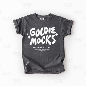 May include: A charcoal gray t-shirt with rolled-up sleeves. The shirt features the white text "GOLDIE MOCKS" in a playful font, with the words "MOCKUP STUDIO" and additional text below. The shirt is a casual wear item.