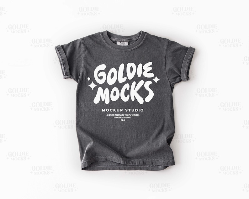 May include: A charcoal gray t-shirt with rolled-up sleeves. The shirt features the white text "GOLDIE MOCKS" in a playful font, with the words "MOCKUP STUDIO" and additional text below. The shirt is a casual wear item.