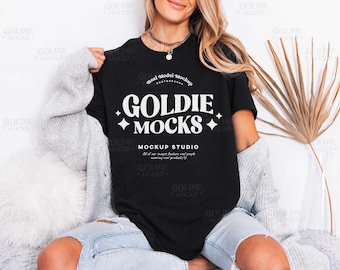 May include: Black t-shirt with the text "GOLDIE MOCKS" in white, with the words "MOCKUP STUDIO" below. The shirt is worn with light blue jeans and a gray cardigan. A necklace is visible.