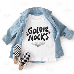 Puede incluir: Camiseta blanca con un gráfico en blanco y negro que dice "Goldie Mocks Mockup Studio". La camiseta está colocada sobre una superficie blanca con una camisa vaquera azul y zapatos negros y blancos de tela.