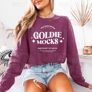 May include: A maroon long-sleeve sweatshirt with the text "GOLDIE MOCKS" in white. The sweatshirt is worn with denim shorts. The image is a mockup for apparel.