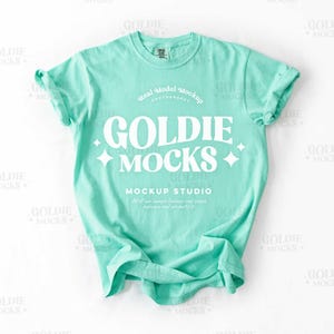 Könnte beinhalten: Ein blaugrünes T-Shirt-Mockup mit dem Text "GOLDIE MOCKS" in weißer Retro-Schrift. Der Text ist von weißen Sternen umgeben. Der Text "Real Model Mockup" und "MOCKUP STUDIO" ist ebenfalls in weißer Schrift sichtbar.