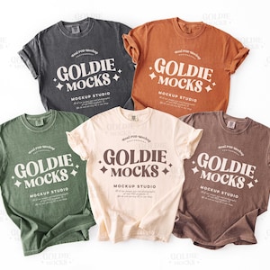 May include: Five different colored t-shirts with the text "Goldie Mocks" and "Mockup Studio" printed on them. The shirts are laid out on a white surface.