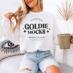 Könnte beinhalten: Ein weißes Sweatshirt mit dem Text "GOLDIE MOCKS" in Schwarz. Die Person hält einen grünen Becher mit dem Wort "COFFEE" darauf. Die Person trägt eine hellblaue Jeans.