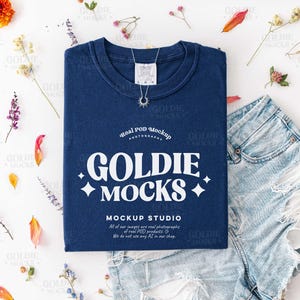 Pode incluir: Uma camiseta azul marinho com o texto "GOLDIE MOCKS" em branco, combinada com calções de ganga. Um colar de prata com um pingente de sol e várias flores e pétalas secas estão dispostas ao redor da camiseta e dos calções em uma superfície branca.
