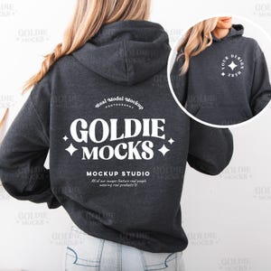 May include: A dark gray hooded sweatshirt with a white logo that says "Goldie Mocks" and "Mockup Studio". The logo is in a retro font style. The sweatshirt is being worn by a person with long brown hair.
