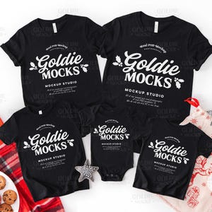 May include: Black t-shirts and a onesie with the text "Goldie Mocks" in white script. The shirts are arranged with holiday-themed decorations, including a star ornament and a Christmas stocking.