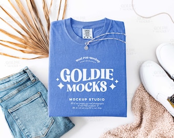 May include: A blue t-shirt with the text "GOLDIE MOCKS" in white, along with a silver necklace, blue jeans, and white sneakers. A dried palm leaf and a beige blanket are also present. The shirt has a tag that says "COMFORT COLORS".