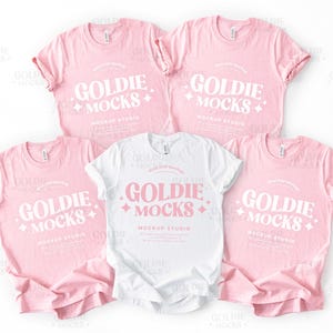 Könnte beinhalten: Fünf T-Shirts, vier rosa und eines weiß, werden gezeigt. Jedes Shirt hat den Text "GOLDIE MOCKS MOCKUP STUDIO" in einer stilisierten Schriftart. Die rosa Shirts haben hochgekrempelte Ärmel. Die Shirts sind auf weißem Hintergrund angeordnet.