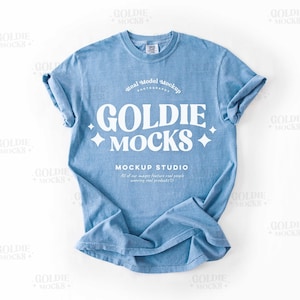 Puede incluir: Una camiseta azul claro con un gráfico blanco que dice "Goldie Mocks Mockup Studio" con un diseño de estrella. La camiseta está arrugada y puesta plana sobre una superficie blanca.
