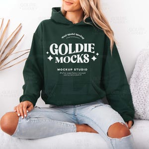 Puede incluir: Una sudadera con capucha verde oscuro con un logotipo blanco que dice "Goldie Mocks" y "Mockup Studio". La sudadera la lleva puesta una persona sentada en una superficie blanca.