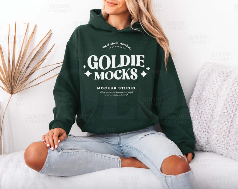 Puede incluir: Una sudadera con capucha verde oscuro con un logotipo blanco que dice "Goldie Mocks" y "Mockup Studio". La sudadera la lleva puesta una persona sentada en una superficie blanca.