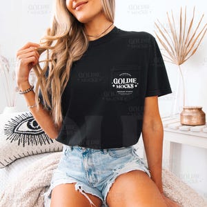 May include: A black cropped t-shirt with a pocket featuring the text "Goldie +Mocks+  Mockup Studio" in white. The shirt is being worn by a person with long blonde hair and denim shorts.