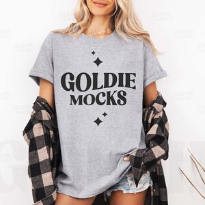 May include: A gray t-shirt with the text "Goldie Mocks" in black lettering. The shirt has a relaxed fit and is styled with a plaid shirt.