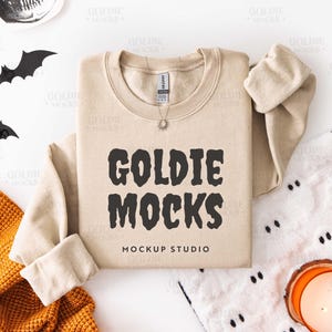 May include: A beige sweatshirt with the text "GOLDIE MOCKS" in black, with "MOCKUP STUDIO" below. The sweatshirt is laid flat with the sleeves folded. Halloween-themed decorations, including bats and a skull, are arranged around the sweatshirt.