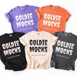 May include: A collection of five t-shirts in various colors: purple, orange, black, cream, and charcoal gray. Each shirt features the text "GOLDIE MOCKS MOCKUP STUDIO" in a bold, white font. The shirts are neatly arranged, showcasing their design.