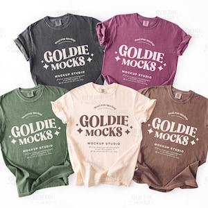 May include: Five different colored t-shirts with the text "Goldie Mocks" and "Mockup Studio" printed on them. The shirts are laid out on a white surface.