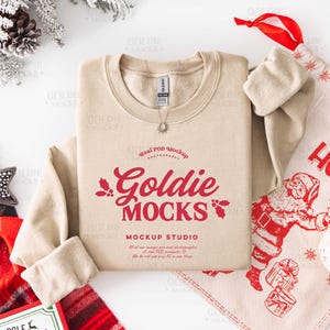 May include: A beige crewneck sweatshirt with the text "Goldie Mocks" in red, along with a Christmas-themed design. A red and black plaid scarf, a star-shaped ornament, and a Santa Claus-themed gift bag are also visible.