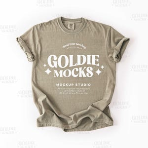 May include: A sage green t-shirt mockup with the text "GOLDIE MOCKS" in white. The text is in a retro font style with a star on each side. The text "Real POD Mockup Photography" is above the main text and the text "MOCKUP STUDIO" is below the main text.