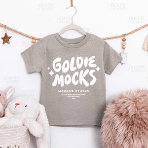 May include: A gray short-sleeve t-shirt with the text "GOLDIE MOCKS" in white, hanging on a hanger. The shirt is part of a mockup studio. Decorative elements include a star, tassels, and stuffed animals.