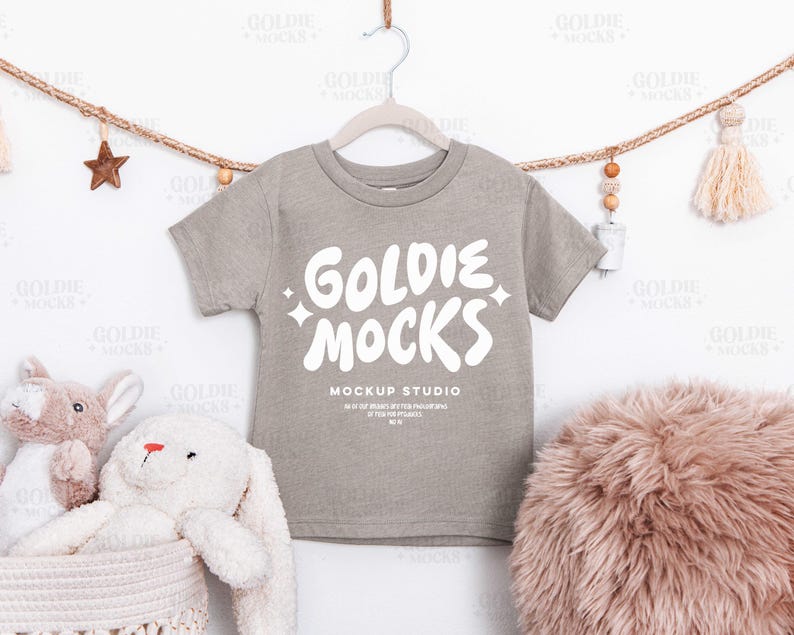 May include: A gray short-sleeve t-shirt with the text "GOLDIE MOCKS" in white, hanging on a hanger. The shirt is part of a mockup studio. Decorative elements include a star, tassels, and stuffed animals.