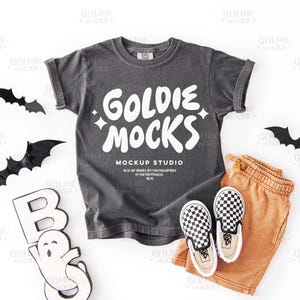 May include: A charcoal grey t-shirt with the text "GOLDIE MOCKS" in white, alongside orange shorts and black and white chequered slip-on shoes. Black bat cutouts and a "BOO" sign are also included.