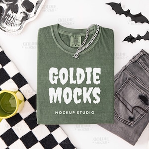 May include: An olive green t-shirt with the text "GOLDIE MOCKS" and "MOCKUP STUDIO" in white. The shirt is paired with gray jeans, a silver chain necklace, and Halloween-themed decorations, including bats and a skull.