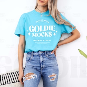 Puede incluir: Una camiseta azul claro con el texto "GOLDIE MOCKS" en blanco, combinada con vaqueros azules rotos. La camiseta tiene cuello redondo y mangas cortas. La imagen es una maqueta de diseño de camiseta.